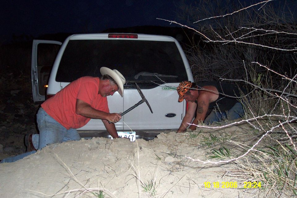 White Ford Excursion rear end buried in sand at night while two men dig it out with a pickaxe