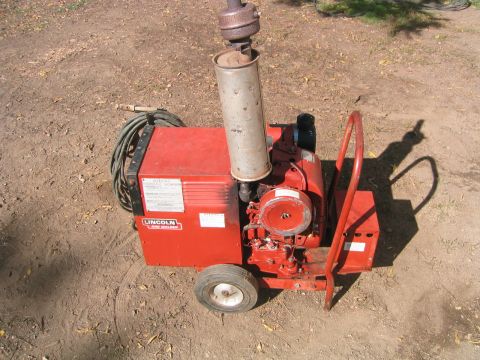 1973 Ford F-100 - A red Lincoln engine-driven arc welder/generator on a two-wheel cart sits on bare dirt, with a tall vertical exhaust stack, air-cooled sing…