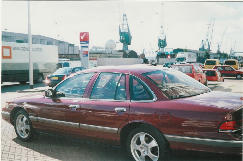 Maroon sedan parked near a harbor with cranes in the background. Other cars and trucks are visible