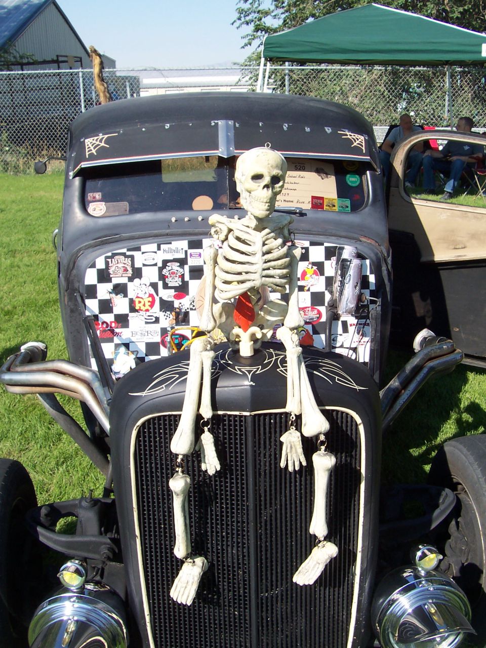 Black hot rod with a skeleton sitting on the hood, checkered flag and stickers on the grille area, and chrome exhaust pipes