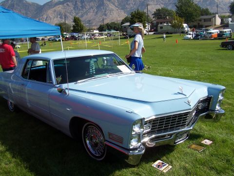 1973 Ford Mustang - This light blue Cadillac Coupe DeVille features a prominent chrome grille and distinctive hood ornament, seen at an outdoor car show.