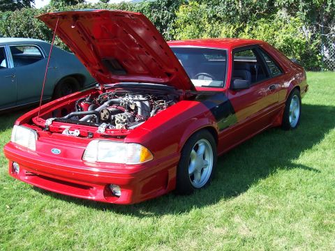 1973 Ford Mustang - This red coupe, likely a 1990s Ford Mustang, is displayed with its hood open, revealing the engine bay and five-star wheels.