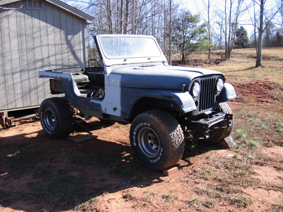 Blue-gray Jeep CJ7 project, doorless and topless, on wood blocks beside a shed with large Sidewinder tires