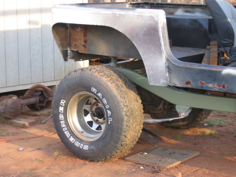 Jeep CJ body with rear fender removed, showing rust and unfinished metal. A spare axle sits nearby