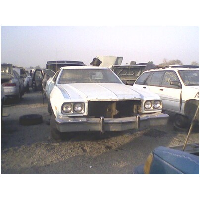 1973 Ford Ranchero - A white 1977 Ford Ranchero sits in a junkyard, its front end damaged and grille missing.
