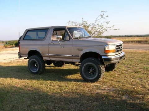 1996 Ford Bronco - Passenger-side profile of a clean tan/mocha Ford Bronco with a matching removable hardtop, parked on dry grass in open country at golden ho…