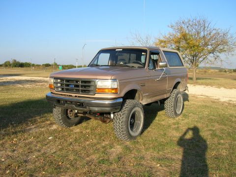 1996 Ford Bronco - Driver-side front three-quarter view of a lifted mocha-tan OBS Ford Bronco parked on grass, showing the body-color grille with blue Ford ov…