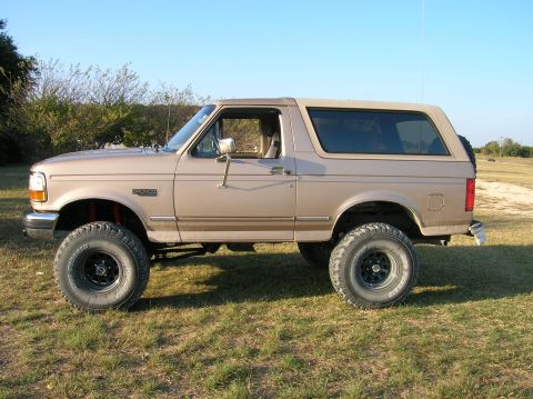 1996 Ford Bronco - Driver-side profile of a lifted tan/mocha OBS Ford Bronco parked on grass, wearing BFGoodrich Mud-Terrain tires on black steel wheels with…