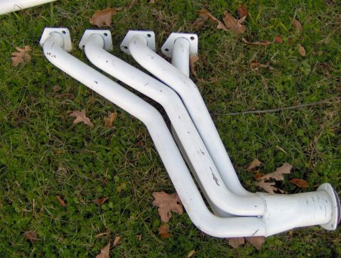 1978 Ford Bronco - A set of white exhaust headers rests on a grassy surface with fallen leaves scattered around.