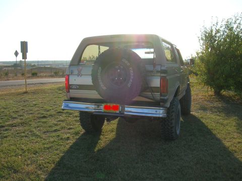 1996 Ford Bronco - Rear view of a lifted tan OBS Ford Bronco backlit by low sun on a grassy roadside, with a Pro Comp Mud Terrain spare tire on a swing-out-st…