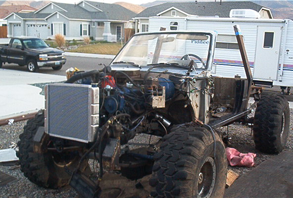 Off-road vehicle chassis with exposed engine and large tires, parked next to a trailer