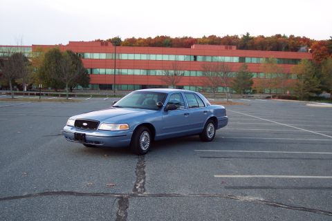2001 Ford Crown Victoria - A blue 2001 Ford Crown Victoria sedan is parked in a large, empty parking lot with a red brick building in the background.