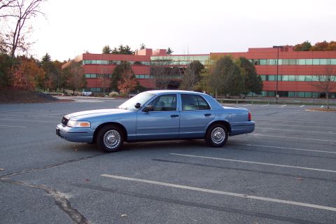2001 Ford Crown Victoria - A blue 2001 Ford Crown Victoria is parked in an empty lot, with a large red office building in the background.