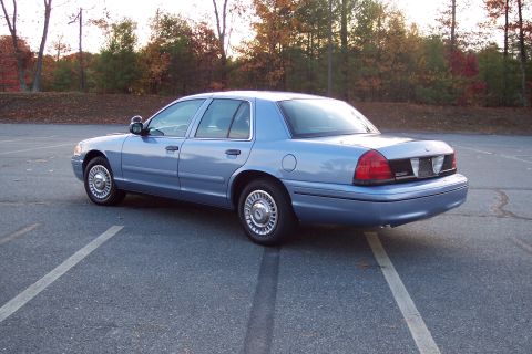 2001 Ford Crown Victoria - A blue 2001 Ford Crown Victoria is parked in a lot with trees in the background.