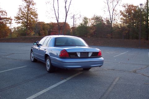 2001 Ford Crown Victoria - A light blue 2001 Ford Crown Victoria sedan is viewed from the rear in a parking lot with autumn trees in the background.