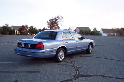 2001 Ford Crown Victoria - A light blue sedan is parked in an empty lot with houses and trees in the background.