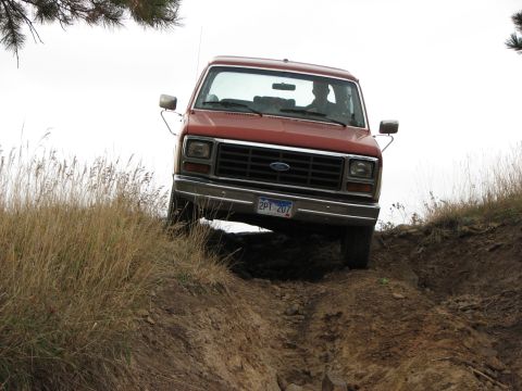 1986 Ford Bronco - A red Ford truck navigates a steep dirt incline, showing its front grille and license plate.