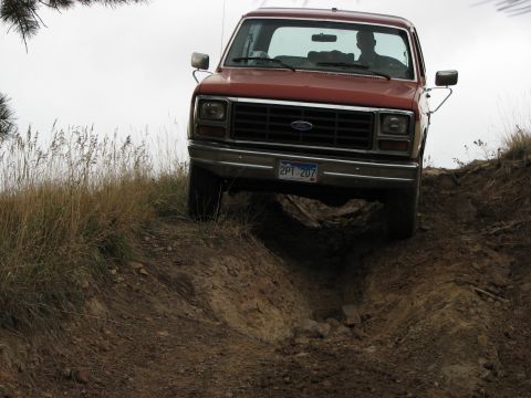 1986 Ford Bronco - A red Ford truck navigates a challenging dirt trail, its front grille and headlights clearly visible.