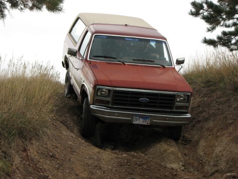 1986 Ford Bronco - 1986 Ford Bronco navigating an off-road trail with dry grass and trees visible.