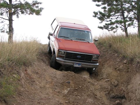 1986 Ford Bronco - This red pickup truck with a camper shell is navigating a steep, rutted dirt path.