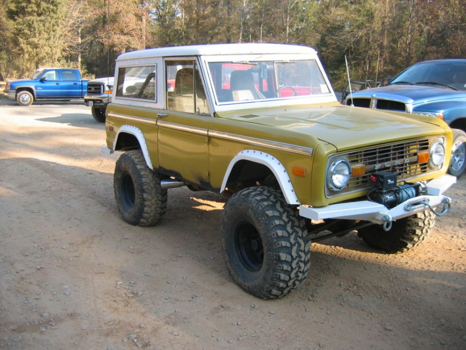 Yellow Ford Bronco with white top and large off-road tires, front bumper with winch. Blue pickup truck in background