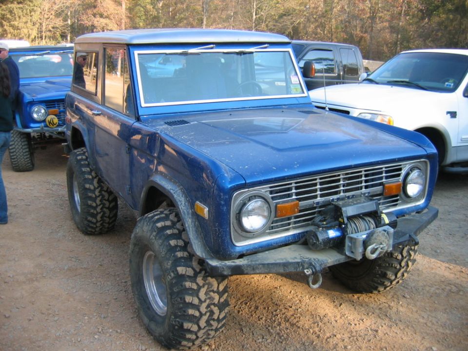 Blue Ford Bronco with large tires and a winch on the front bumper. Another blue Bronco is visible behind it