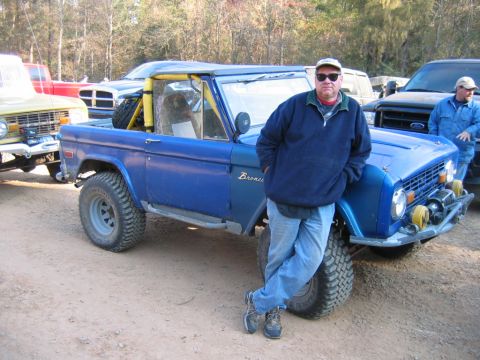 1992 Ford Mustang - A man leans against a blue Ford Bronco, which has a roll cage and off-road tires. The word "Bronco" is visible on the side.