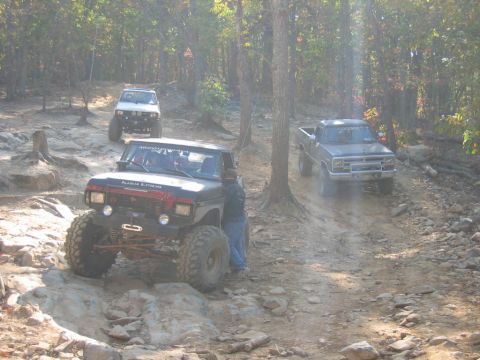 1992 Ford Mustang - Multiple trucks navigate a challenging off-road course in a forest setting.