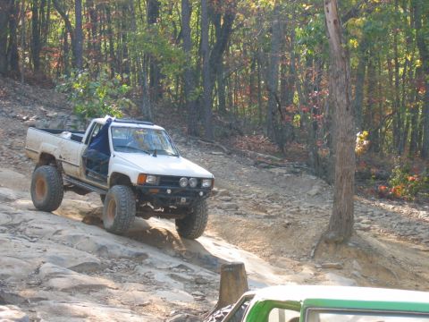 1992 Ford Mustang - Modified Toyota pickup truck navigating a challenging off-road trail with wooded scenery.