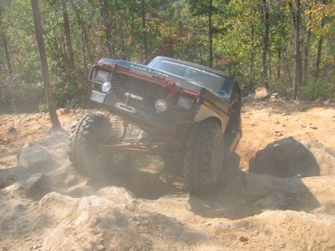 1992 Ford Mustang - This heavily modified truck is navigating a challenging off-road course with large rocks and trees in the background.
