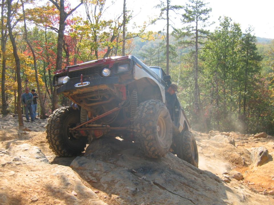 Off-road vehicle with large tires climbing a rock face, red suspension, and a winch on the front bumper