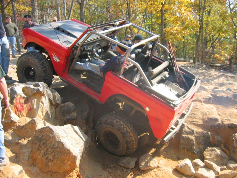 Red off-road vehicle with roll cage climbing over large rocks on a dirt trail. The vehicle has large tires and a modified hood