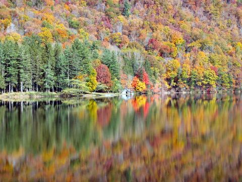 1985 Ford LTD Crown Victoria - A lake reflects the vibrant colors of autumn trees lining the shore, with a small boathouse visible amongst the foliage.