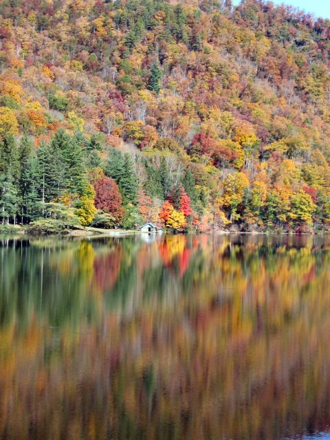 1985 Ford LTD Crown Victoria - A lake reflects the vibrant colors of autumn trees blanketing the hillside, with a small boathouse visible on the shore.