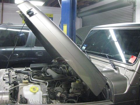 1987 Suzuki Samurai - The engine compartment of a silver Jeep Wrangler is visible with its hood propped open, while another SUV is parked behind it.