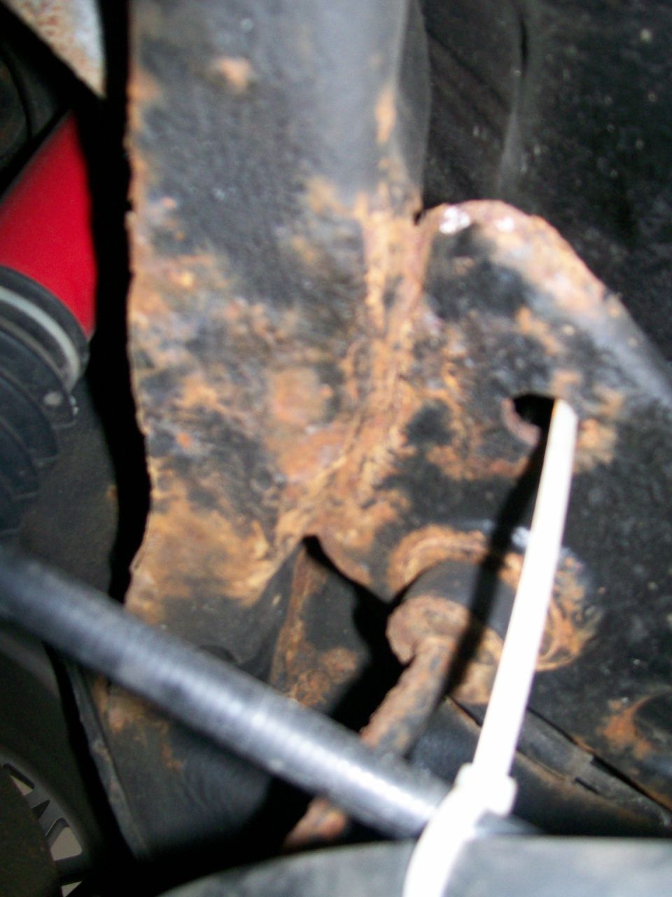 Rusted metal suspension part with a red hose and black tubing, secured by a white zip tie