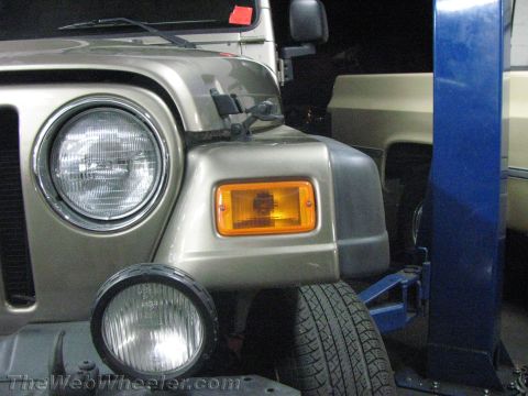 1987 Suzuki Samurai - Close-up of a Jeep Wrangler's front end, showing its headlight and turn signal assembly. A second vehicle and a lift are also in view.