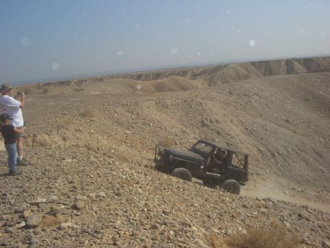 1985 Ford Bronco - A Jeep navigates a steep, rocky incline in a desert landscape, while two onlookers observe from the side.