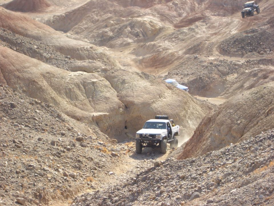 White off-road truck driving down a rocky, barren canyon with two other vehicles visible in the distance