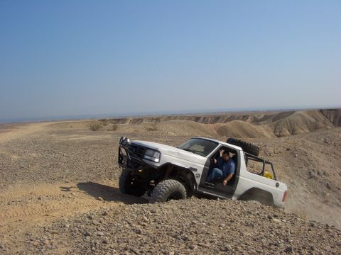 1985 Ford Bronco - A white off-road SUV navigates a steep, rocky terrain under a clear blue sky.