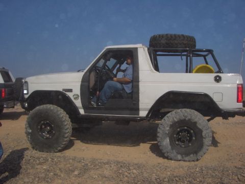 1985 Ford Bronco - Modified 1985 Ford Bronco with off-road tires and roll cage, parked on a dirt surface.
