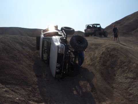 1985 Ford Bronco - A white truck has rolled over on a steep dirt incline, with another vehicle and a person observing the scene.