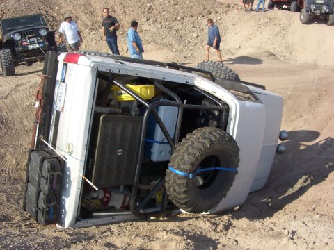1985 Ford Bronco - A white 1985 Ford Bronco lies on its side after an off-road incident, with a spare tire secured to its exterior.
