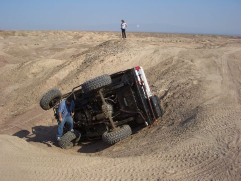 1985 Ford Bronco - A vehicle has rolled over on a sandy incline, with two individuals observing the scene.