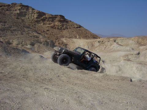 1985 Ford Bronco - A Jeep Wrangler navigates a steep, sandy incline in a barren, rocky desert environment under a clear blue sky.