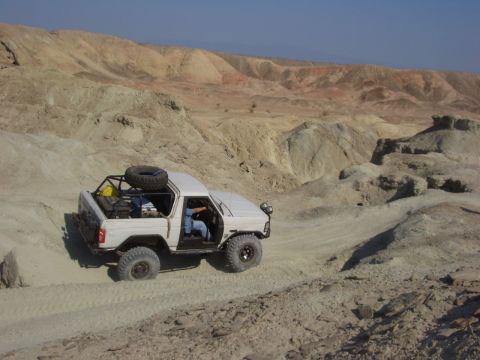 1985 Ford Bronco - A white off-road vehicle navigates a dusty trail through a barren, hilly desert terrain under a clear blue sky.
