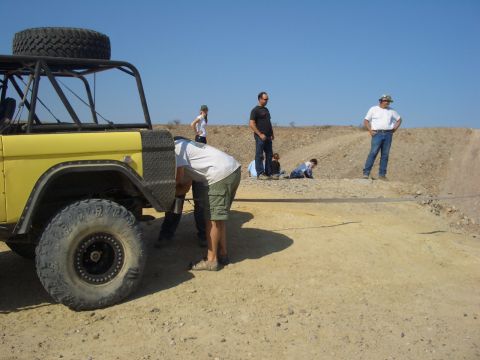 1985 Ford Bronco - A group of people gather around a yellow off-road vehicle in a dry, dusty environment under a clear blue sky.