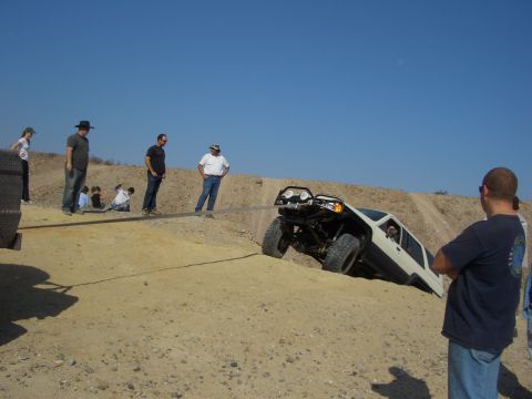 1985 Ford Bronco - An off-road vehicle navigates a steep sandy incline, assisted by a tow rope and onlookers.