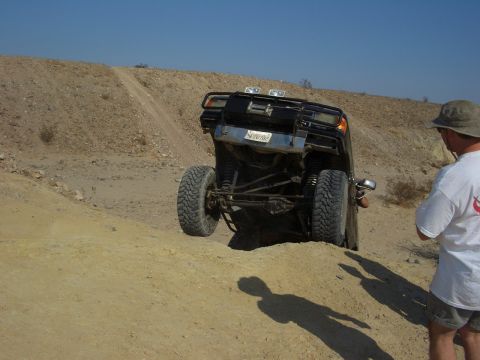 1985 Ford Bronco - A black SUV navigates a steep dirt incline, its rear end angled upwards. A person in a hat watches from the side.