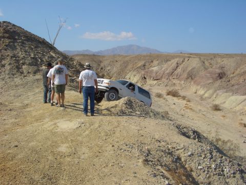 1985 Ford Bronco - A white Ford Bronco is stuck on a steep, dusty incline in a desert landscape with several people observing.